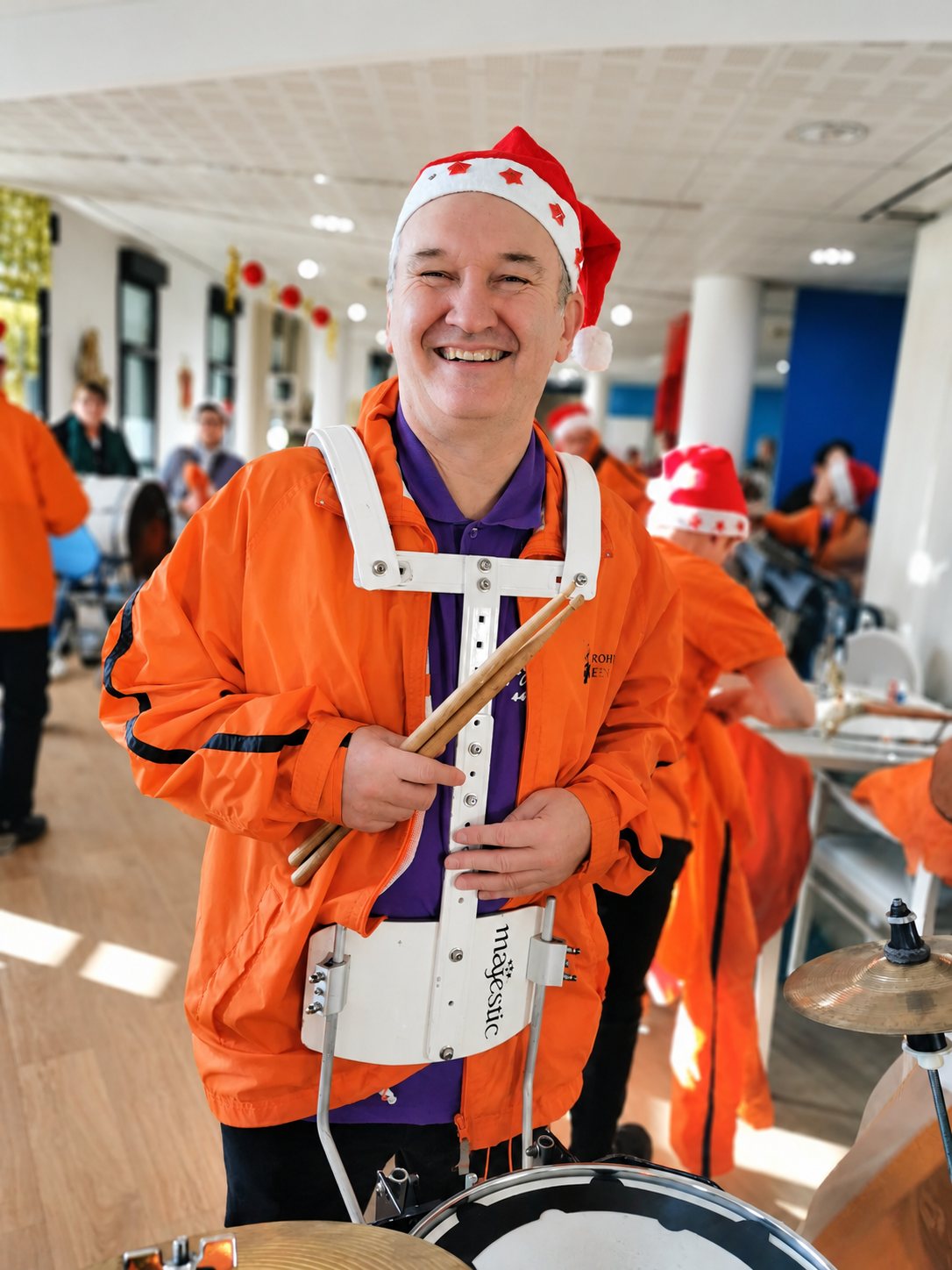 Un homme souriant en veste orange et bonnet de Noël tient des baguettes devant sa caisse claire dans une grande salle décorée.