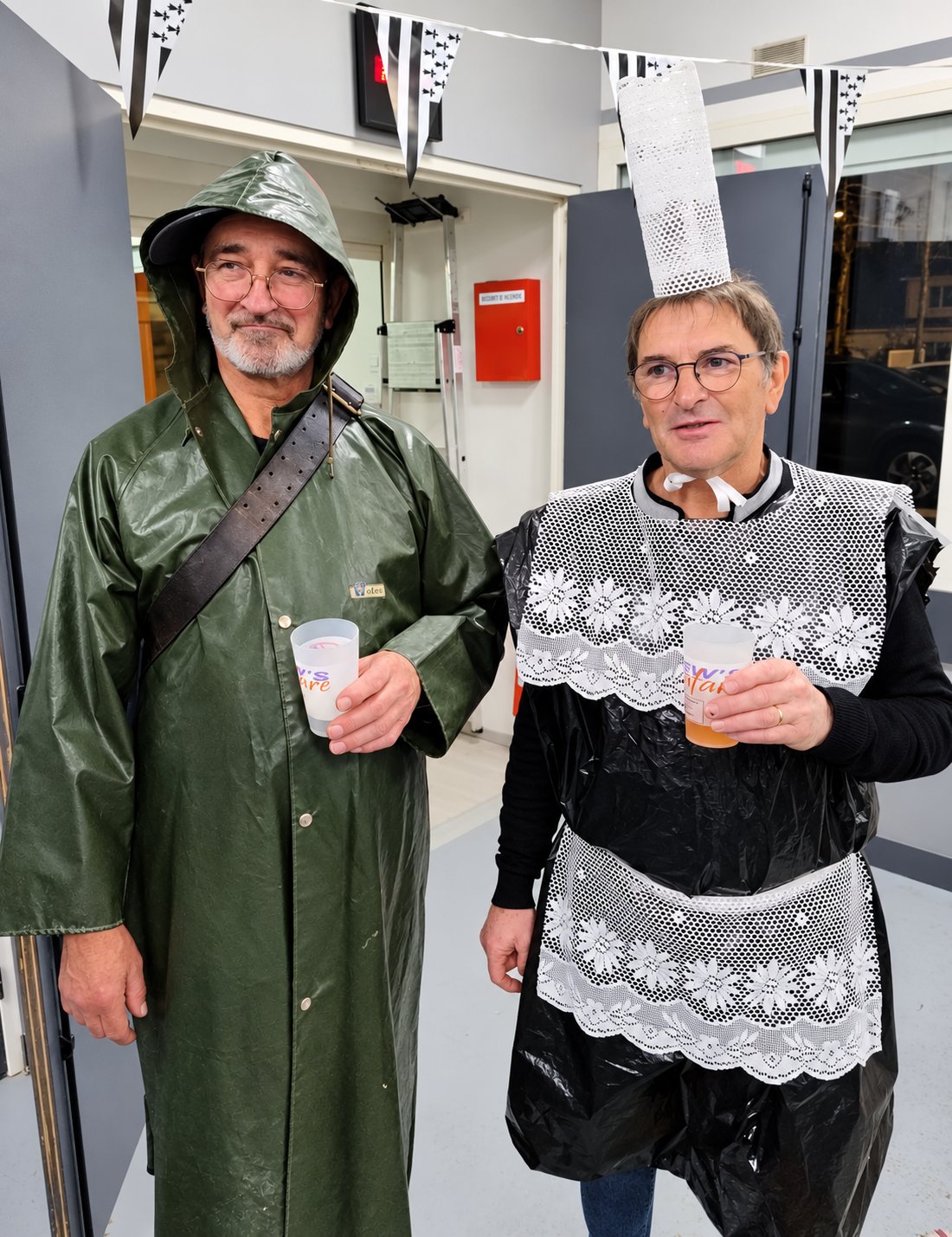 Deux hommes posent en costumes bretons humoristiques, l’un en ciré vert de marin, l’autre avec une coiffe et un tablier en dentelle sur un sac plastique noir, chacun tenant un gobelet.