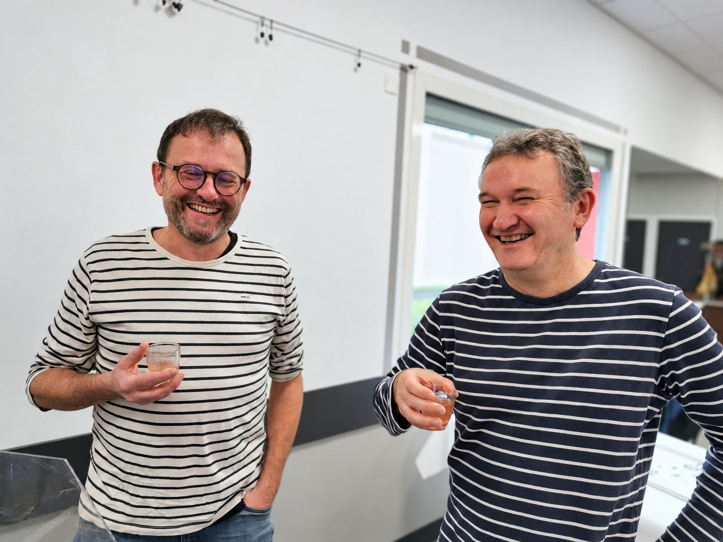 Deux hommes souriants en pulls rayés tiennent chacun un petit verre de dégustation dans une salle aux murs blancs.