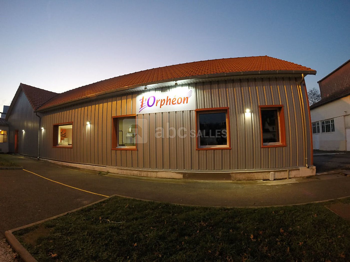 Bâtiment en bardage métallique avec toit en tuiles orange et enseigne L’Orphéon, photographié au crépuscule avec des appliques allumées.