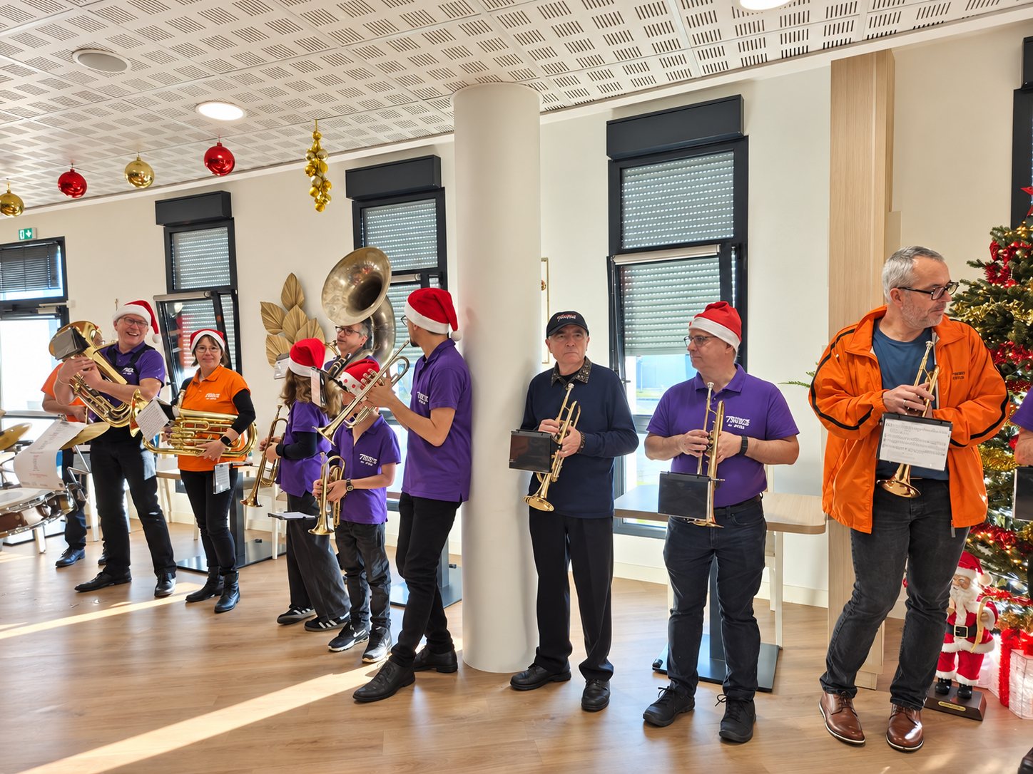 Des musiciens coiffés de bonnets de Père Noël jouent de la trompette, du tuba et du sousaphone dans une salle lumineuse près d’un sapin décoré.