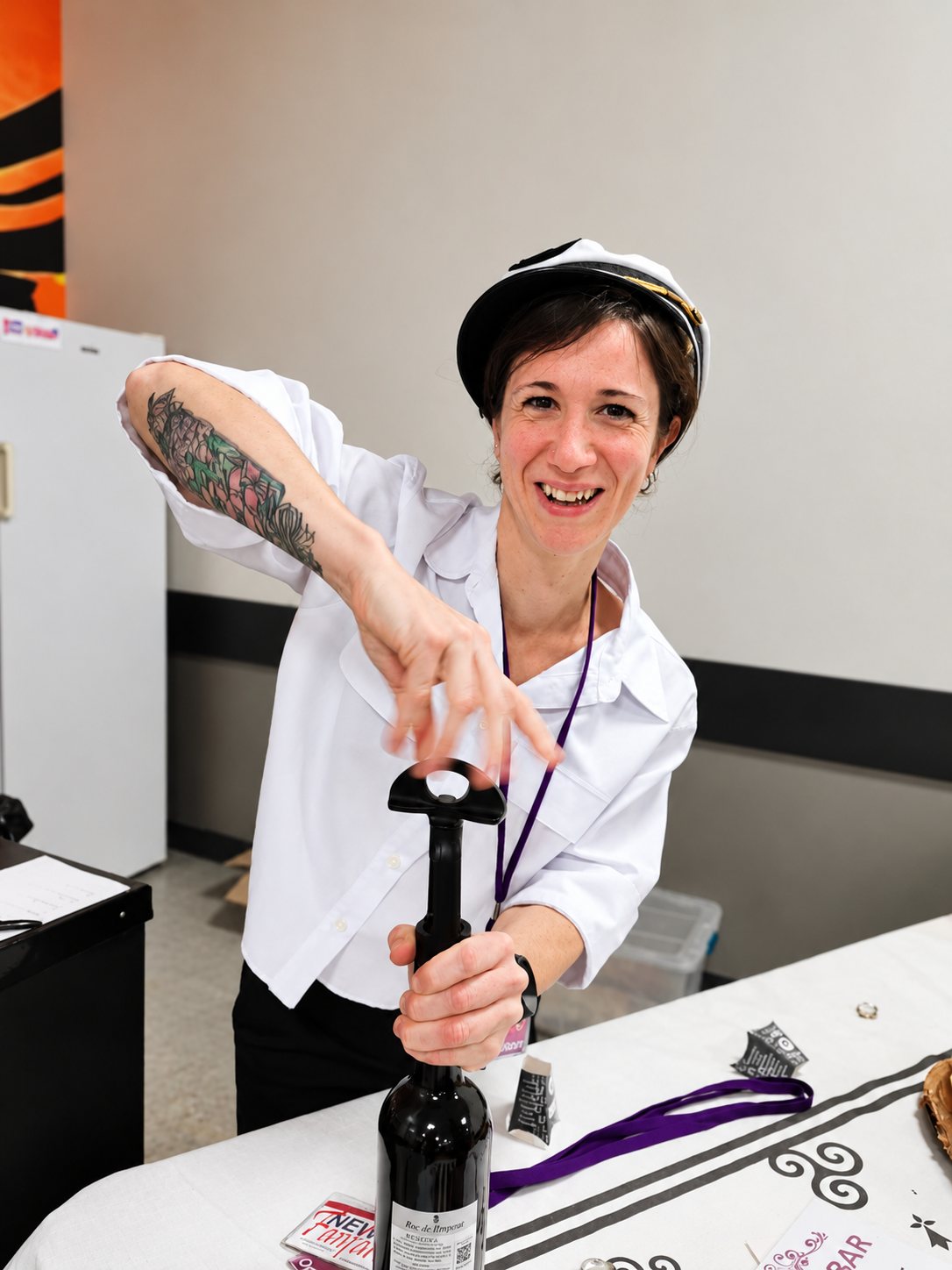 Une femme tatouée, coiffée d’une casquette de marin et souriante, débouche une bouteille de vin avec un tire-bouchon à pompe sur une table de bar.
