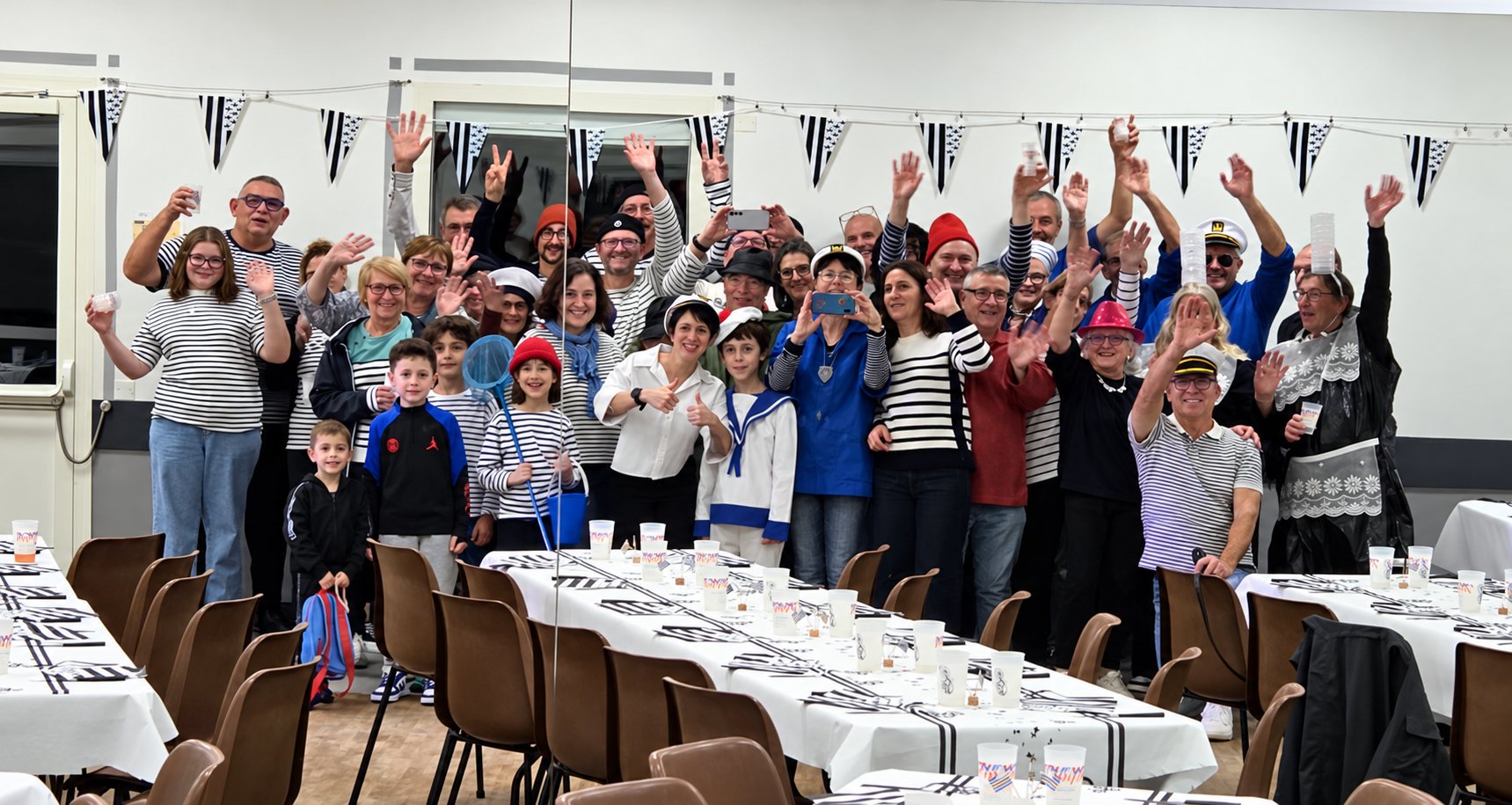 Un grand groupe d’adultes et d’enfants vêtus de rayures marinières lève les bras derrière des tables dressées de blanc, sous des fanions noirs et blancs.