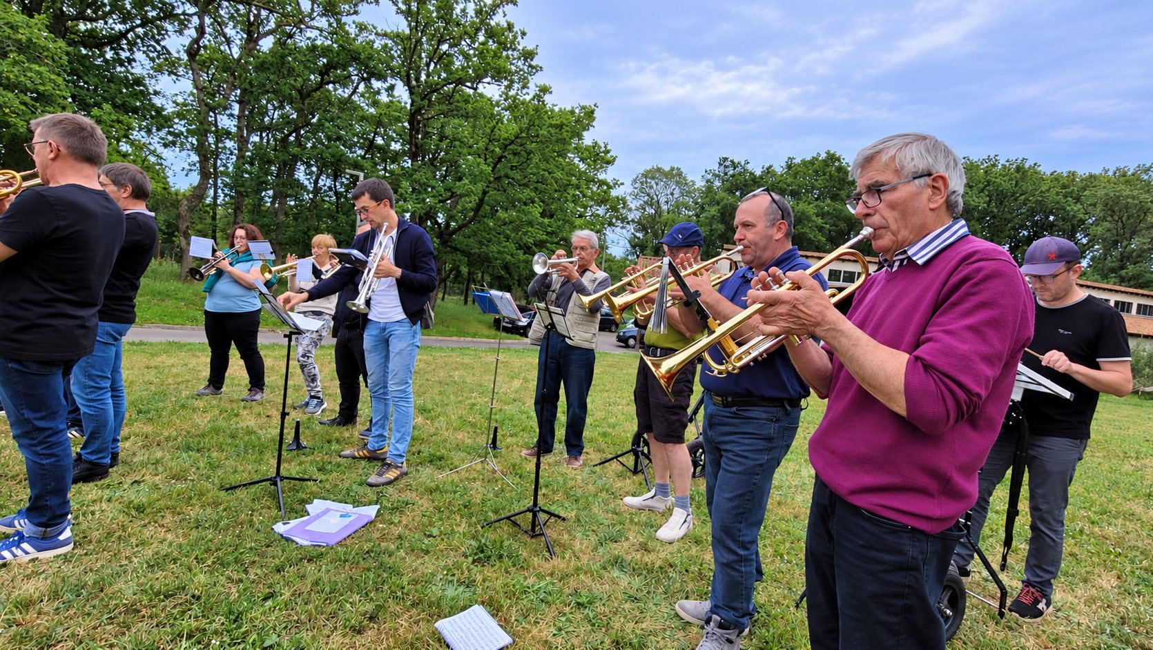 Un groupe de musiciens joue de la trompette sur une pelouse, devant des pupitres, entouré d’arbres sous un ciel nuageux.