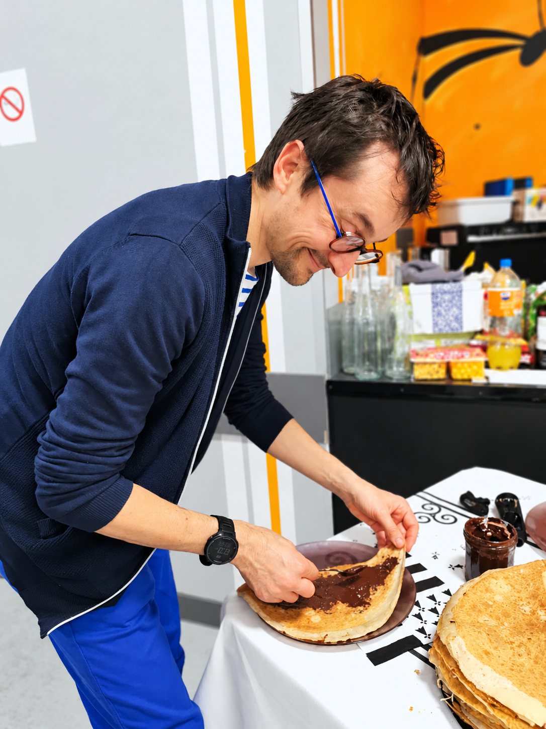 Un homme à lunettes sourit en pliant une crêpe tartinée de chocolat, à côté d’une pile de crêpes sur une table recouverte d’une nappe blanche.
