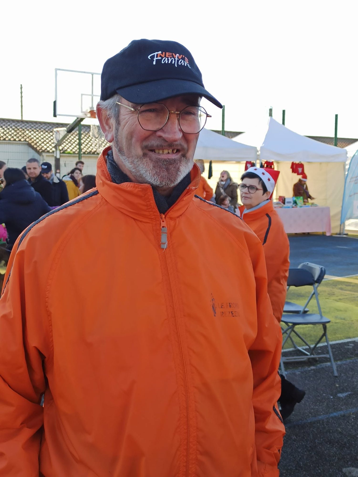 Un homme souriant portant une casquette bleu marine et une veste orange pose devant des tentes blanches et des visiteurs lors d’un marché de Noël en extérieur.
