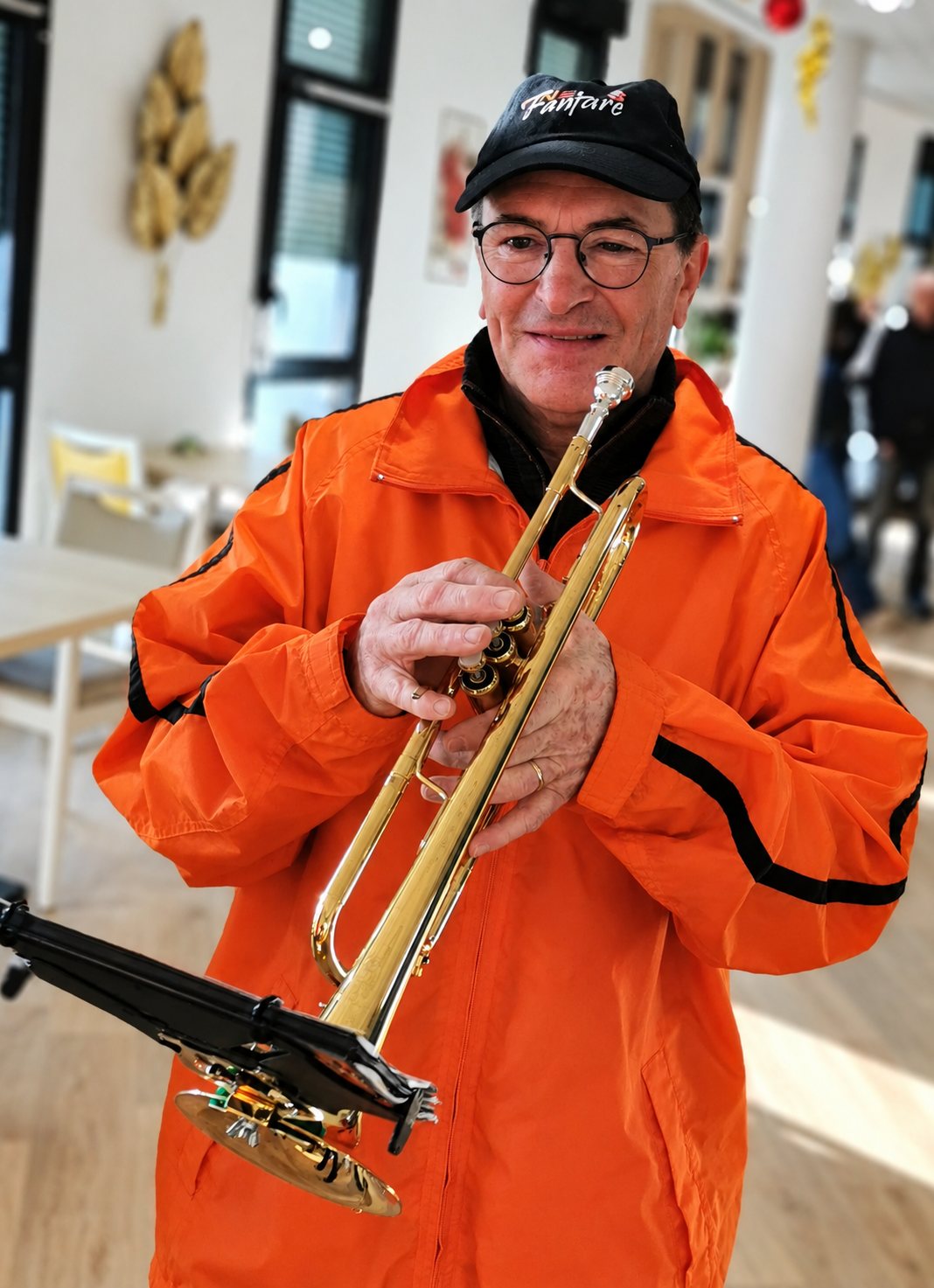 Un homme âgé à lunettes, coiffé d’une casquette noire et vêtu d’une veste orange, sourit en tenant une trompette dorée dans une salle lumineuse.