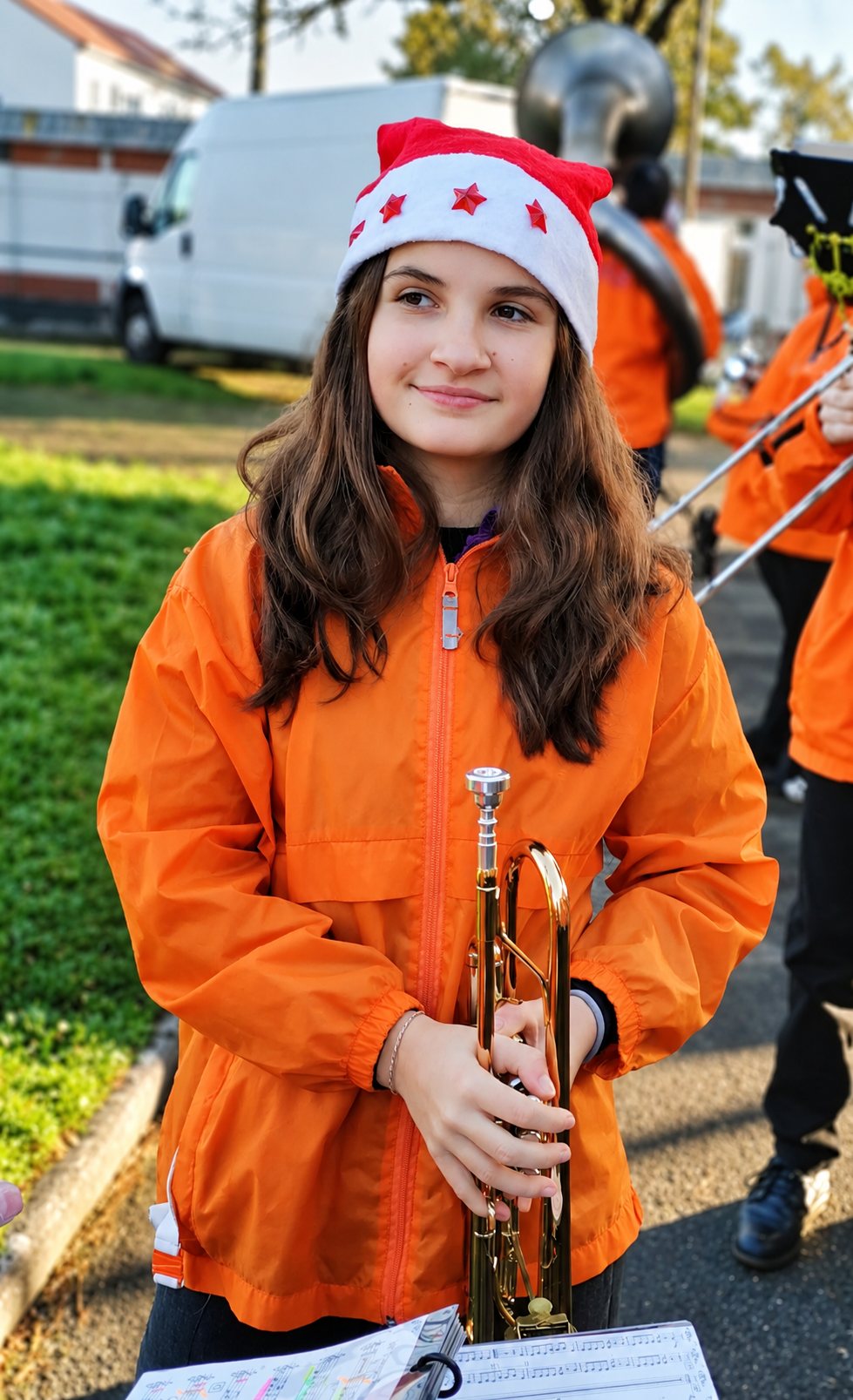 Une jeune fille portant un bonnet de Noël et une veste orange tient une trompette devant une partition, avec d’autres musiciens en orange flous à l’arrière-plan.