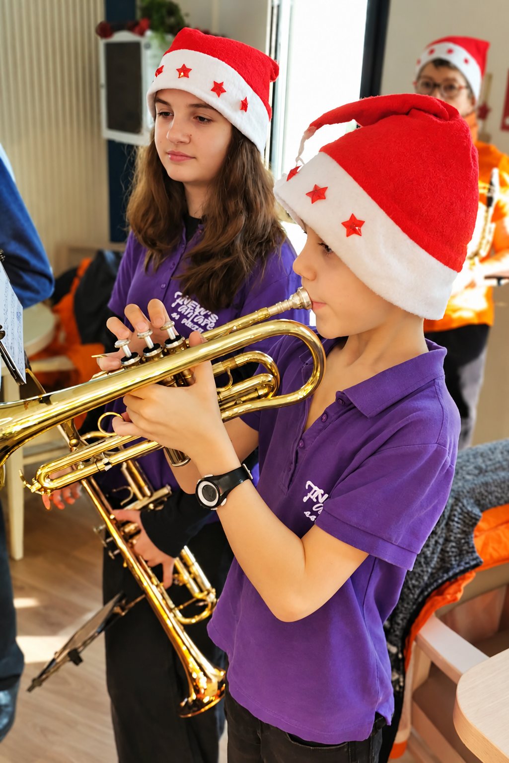 Un garçon en polo violet et bonnet de Père Noël joue de la trompette, à côté d’une jeune fille coiffée du même bonnet.