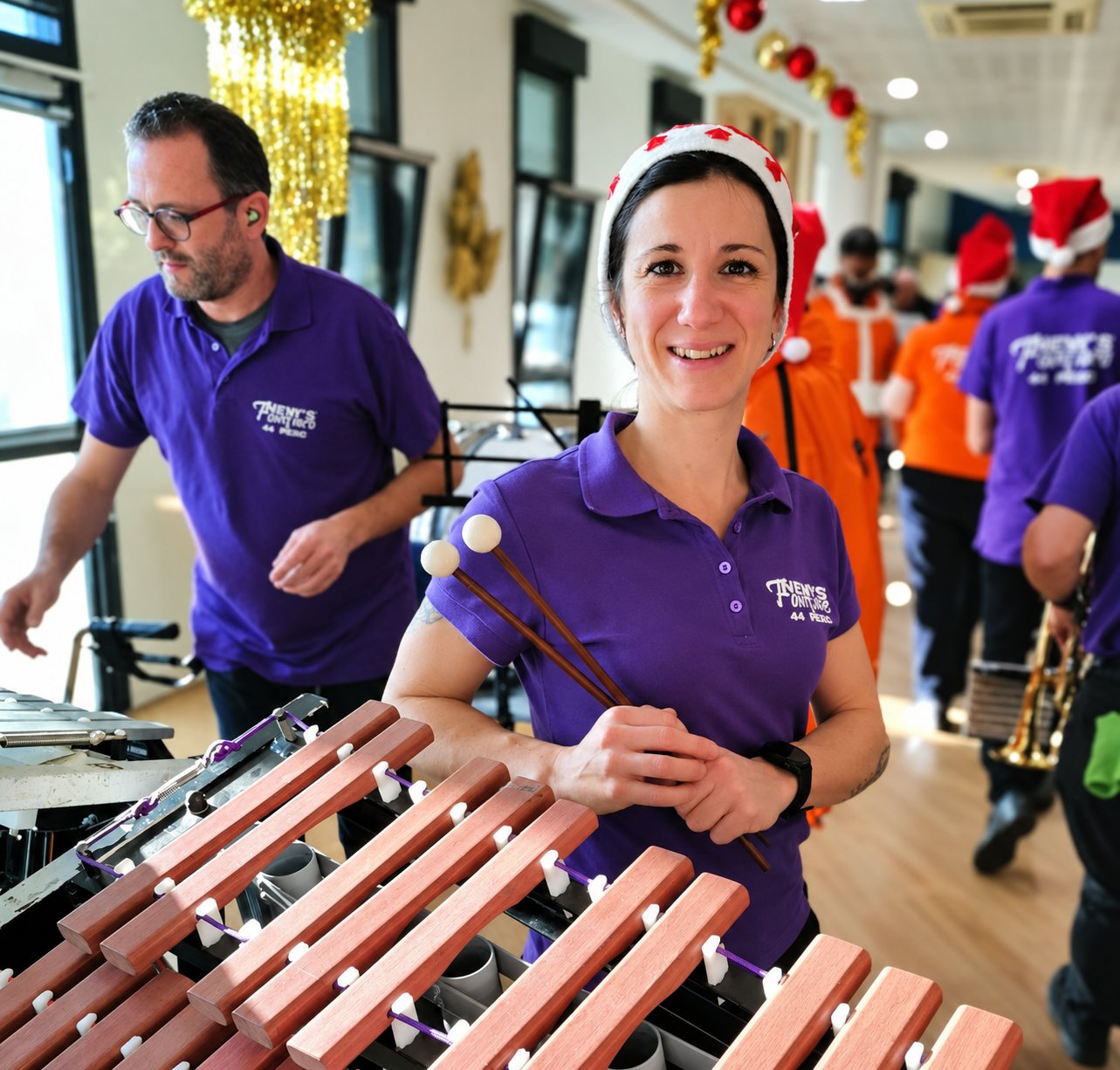 Une musicienne en polo violet et bonnet de Noël sourit en tenant des baguettes derrière un xylophone, entourée des membres de sa fanfare.