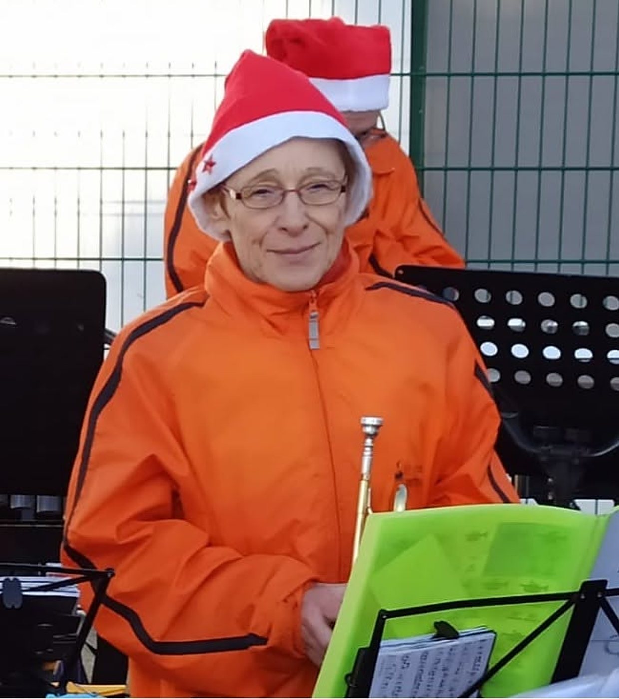 Une musicienne souriante portant une veste orange et un bonnet de Père Noël tient une trompette devant des pupitres, avec un autre musicien coiffé du même bonnet derrière elle.