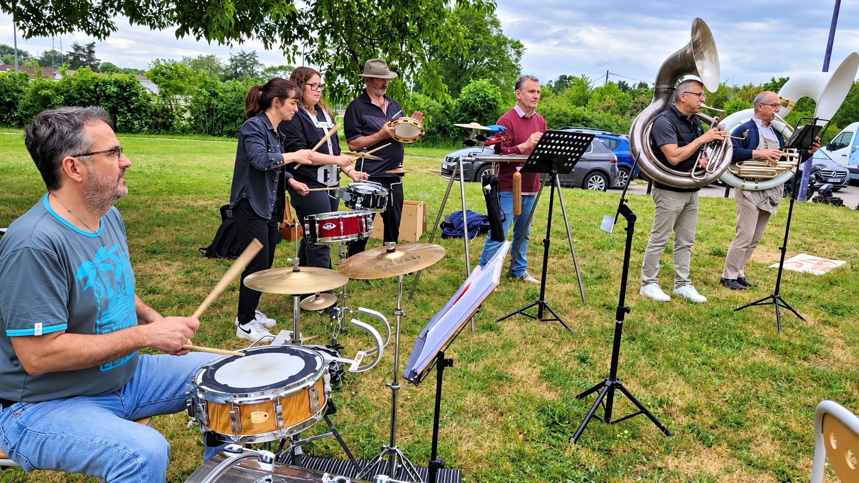 Un groupe de musiciens joue en plein air sur l’herbe, avec un batteur au premier plan, des percussionnistes et deux joueurs de cuivres à droite.