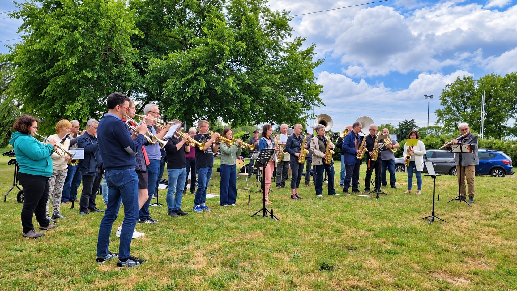 Un groupe de musiciens avec trompettes, saxophones et tubas joue en plein air sur l’herbe, devant de grands arbres et un ciel nuageux.