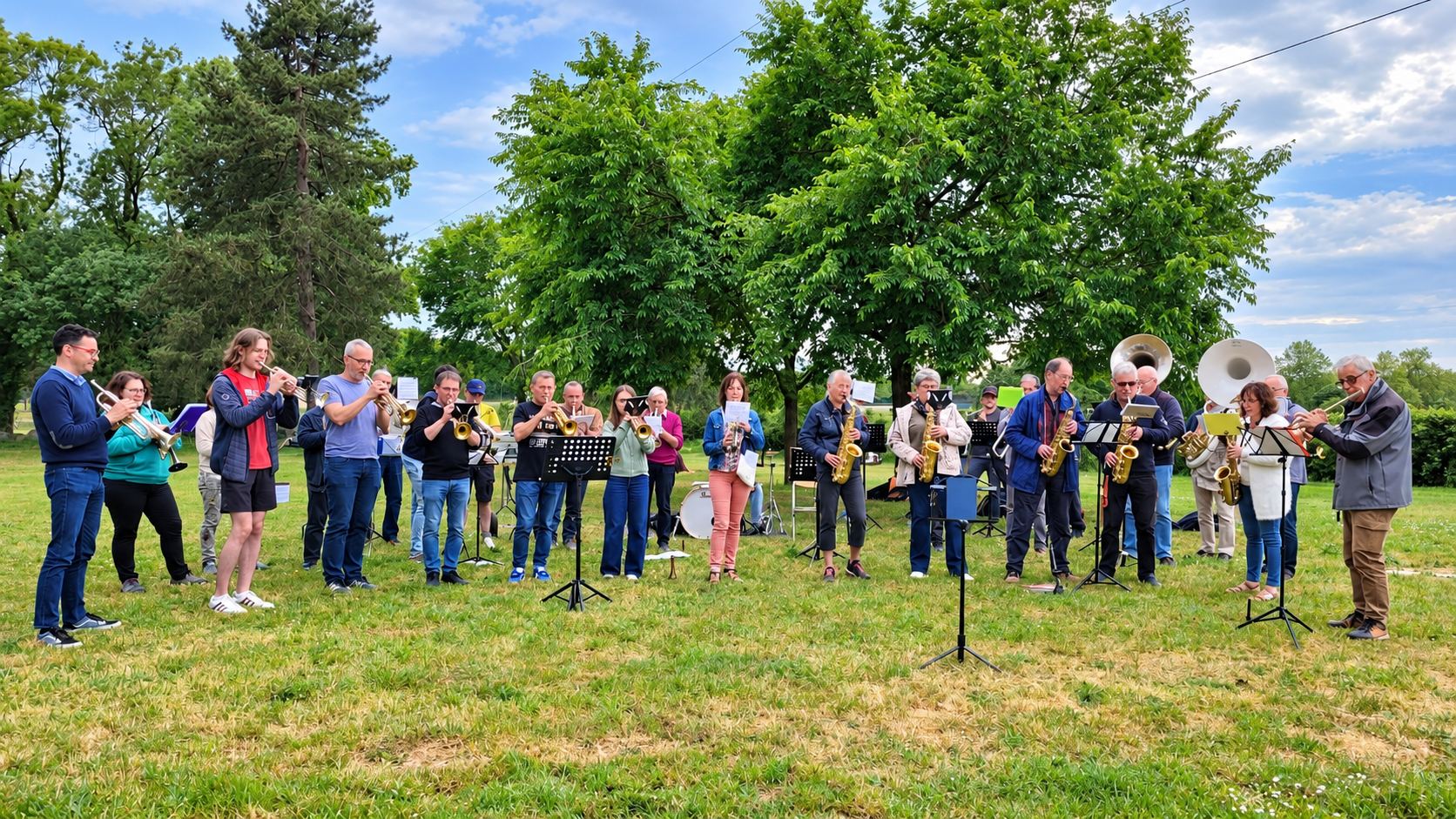 Un groupe de musiciens de tous âges joue de la trompette, du saxophone et du sousaphone sur une pelouse, devant de grands arbres, avec des pupitres devant eux.