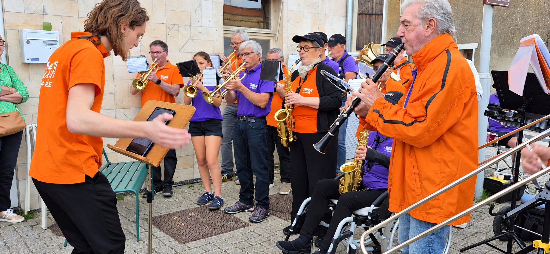 Un chef d’orchestre en t-shirt orange dirige des musiciens en orange et violet, avec trompettes, saxophones et clarinette, lors d’un concert en plein air.