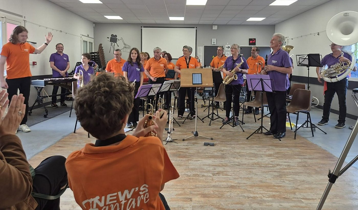 Dans une salle polyvalente, des musiciens en polos orange et violets, avec saxophone, trompettes et sousaphone, se tiennent debout face à un public qui applaudit.