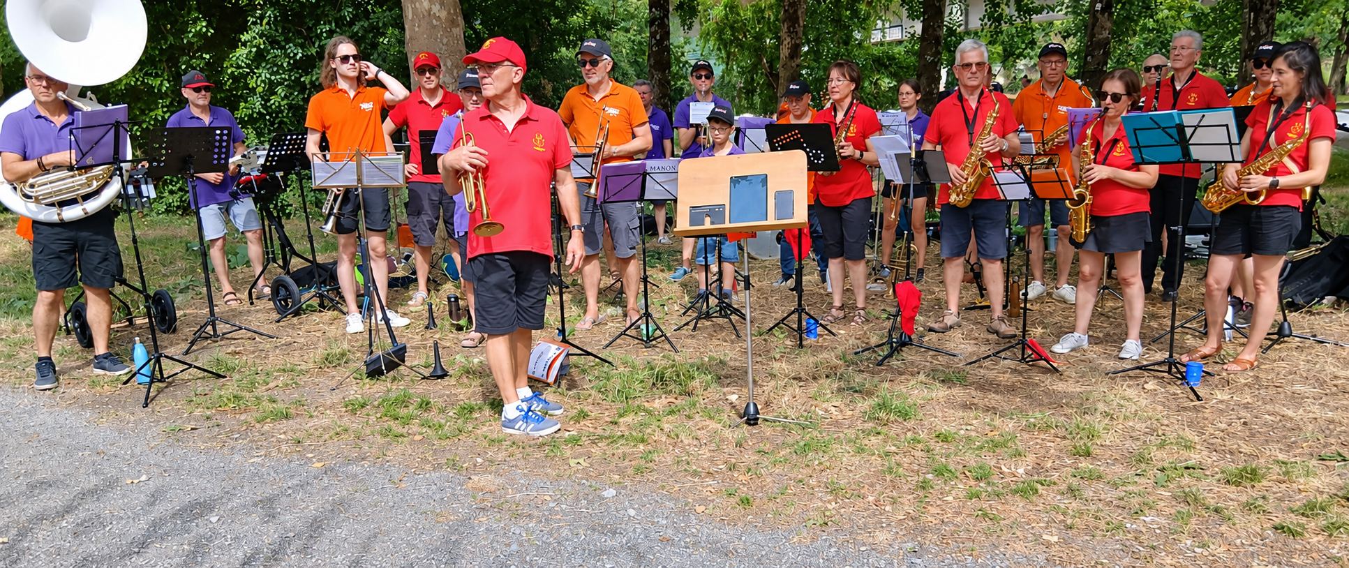 Un groupe de musiciens en polos rouges, oranges et violets joue de la trompette, du saxophone et du sousaphone sur un terrain herbeux sous les arbres, avec des pupitres devant eux.