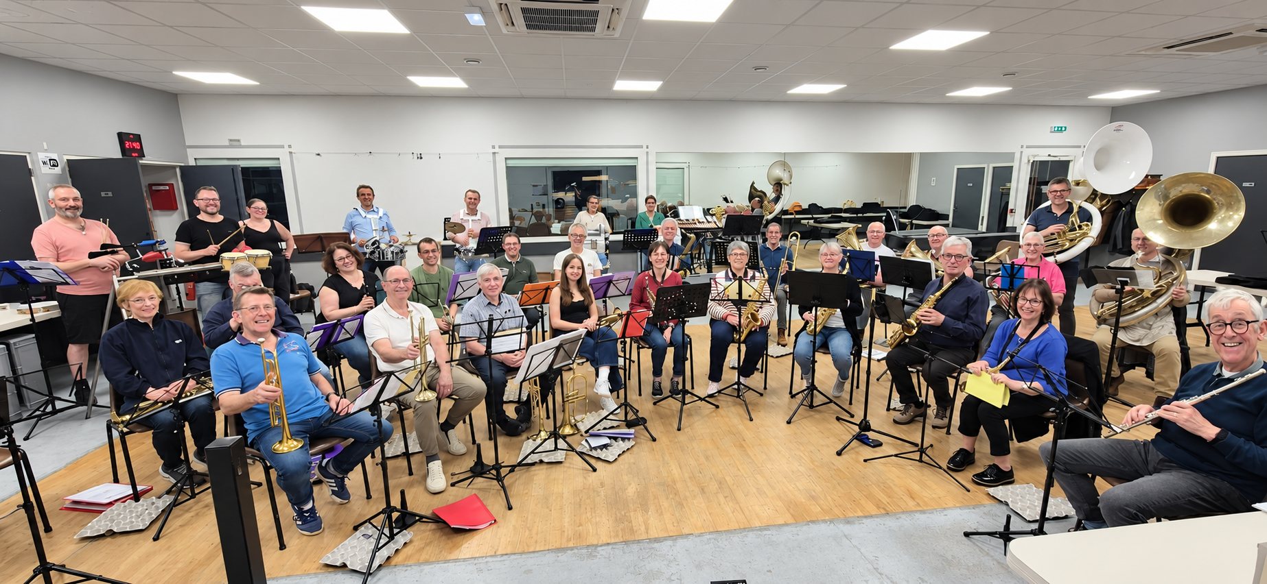 Un groupe de musiciens d’un orchestre d’harmonie pose avec trompettes, saxophones, flûtes, sousaphones et percussions dans une salle de répétition avec pupitres.