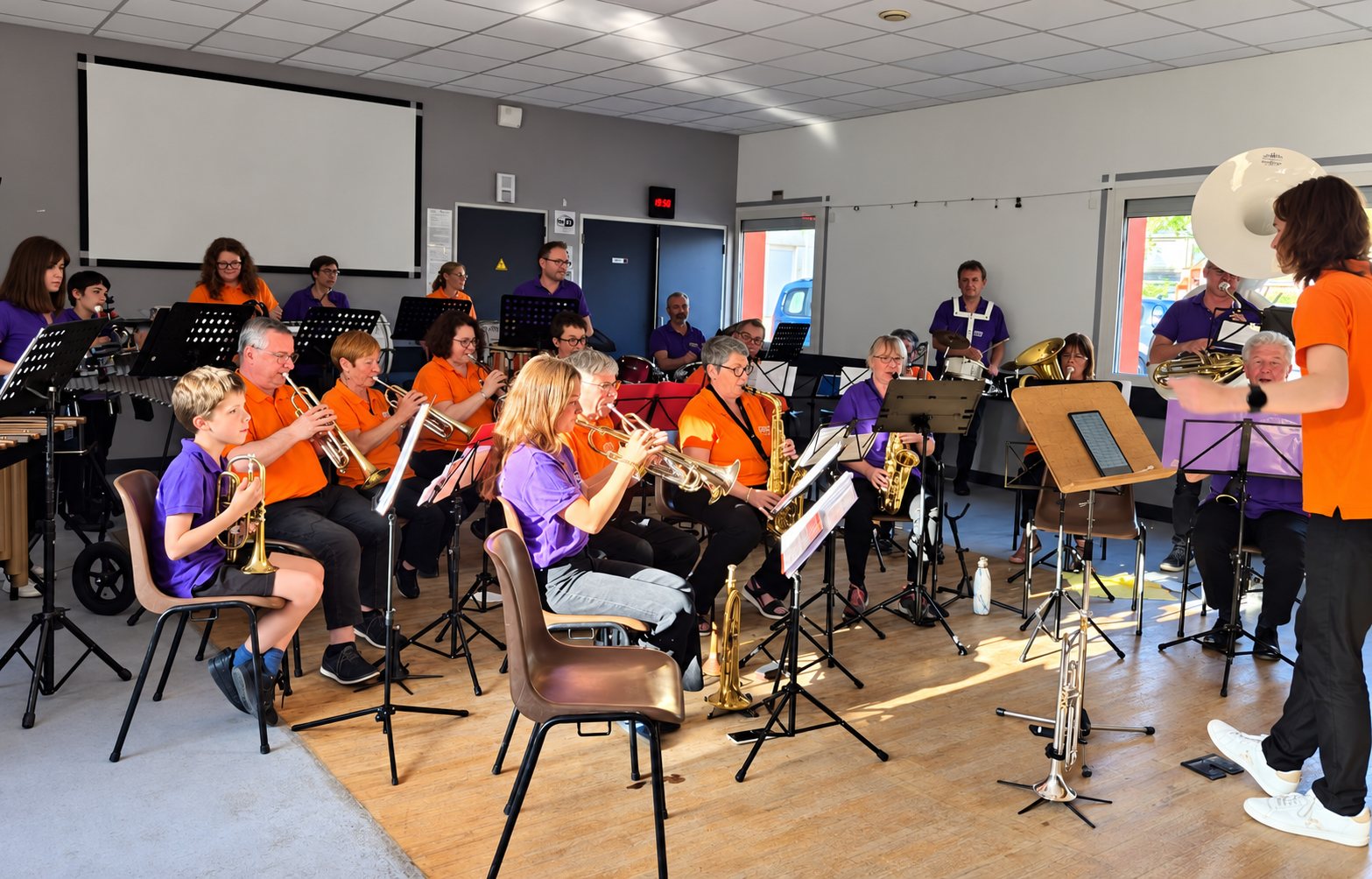 Des musiciens vêtus d’orange et de violet jouent de la trompette, du saxophone et du sousaphone dans une salle pendant une répétition dirigée par un chef.