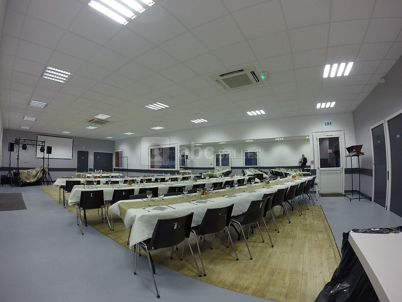 Grande salle de réception au sol gris et parquet clair, équipée de longues tables aux nappes blanches et de chaises noires, avec un écran et du matériel de sonorisation au fond.
