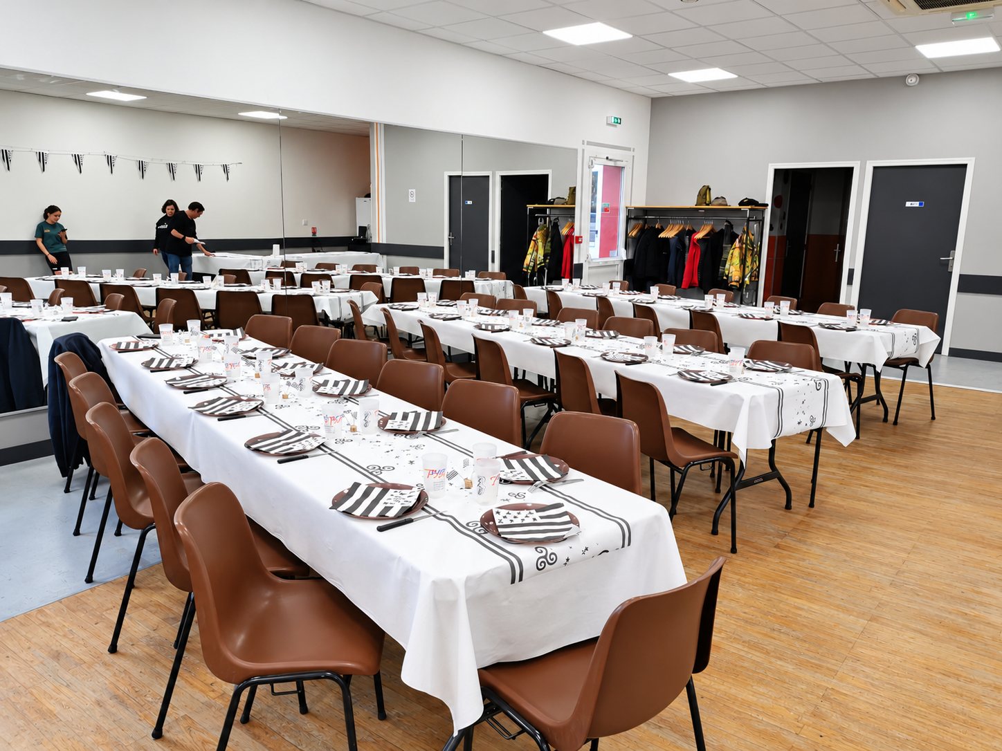 Une grande salle lumineuse avec de longues tables couvertes de nappes blanches, d’assiettes à motif rayé noir et blanc et de chaises marron, prête pour un repas.