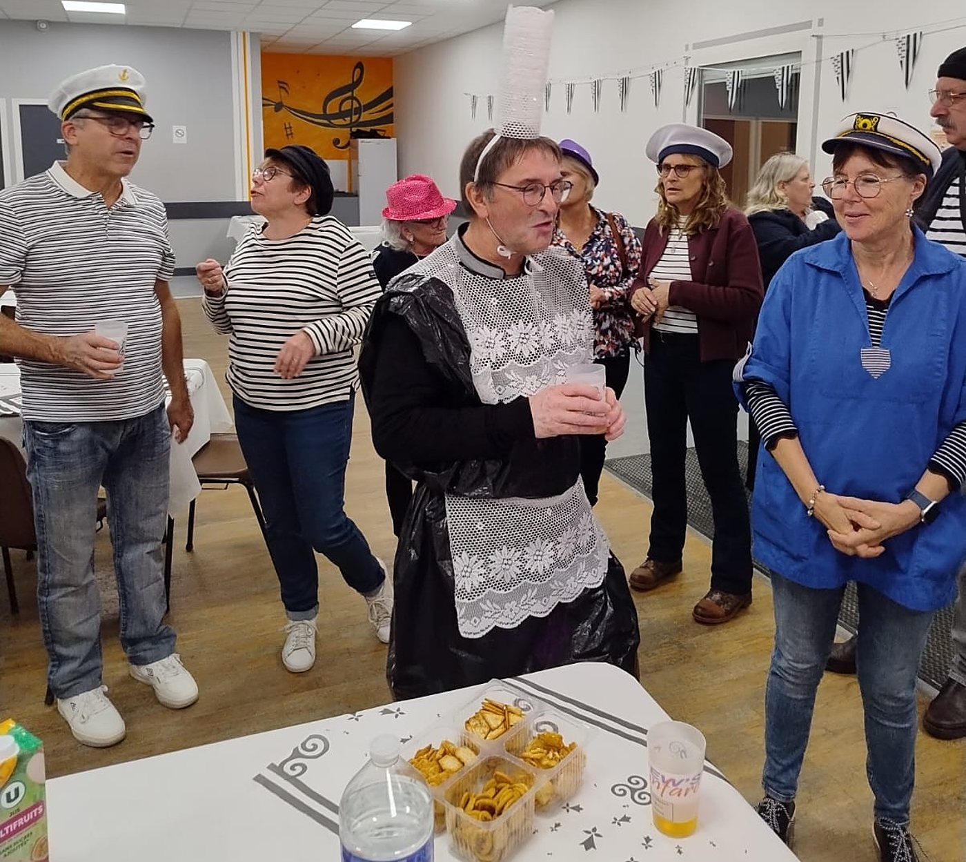 Des personnes déguisées en marins et en Bretonne avec une coiffe en gobelets bavardent autour d’une table garnie de biscuits apéritifs et de boissons.