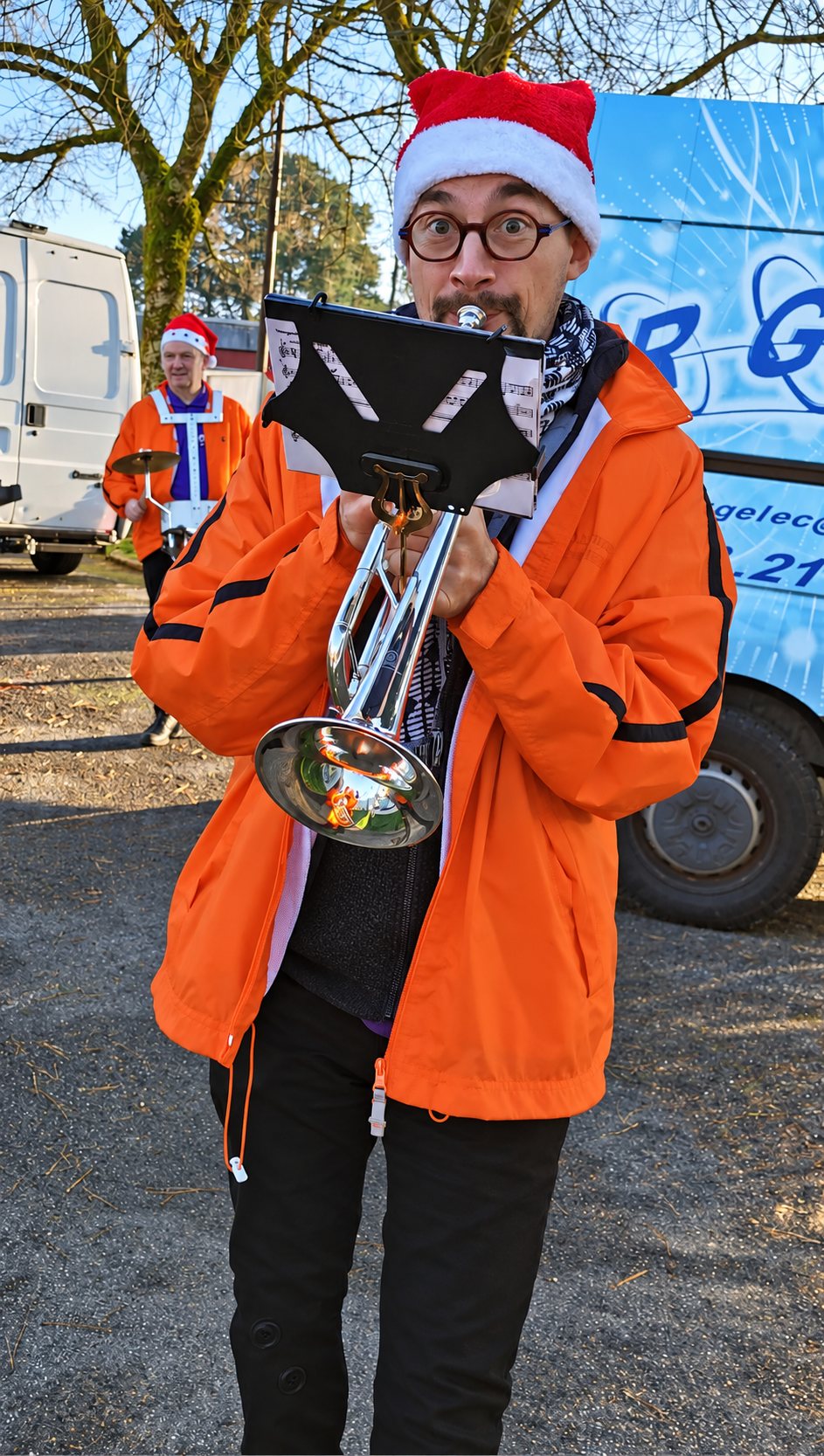 Un musicien portant un bonnet de Père Noël et une veste orange joue de la trompette avec une partition fixée, tandis qu’un autre musicien avec une cymbale se tient derrière lui près de camionnettes.